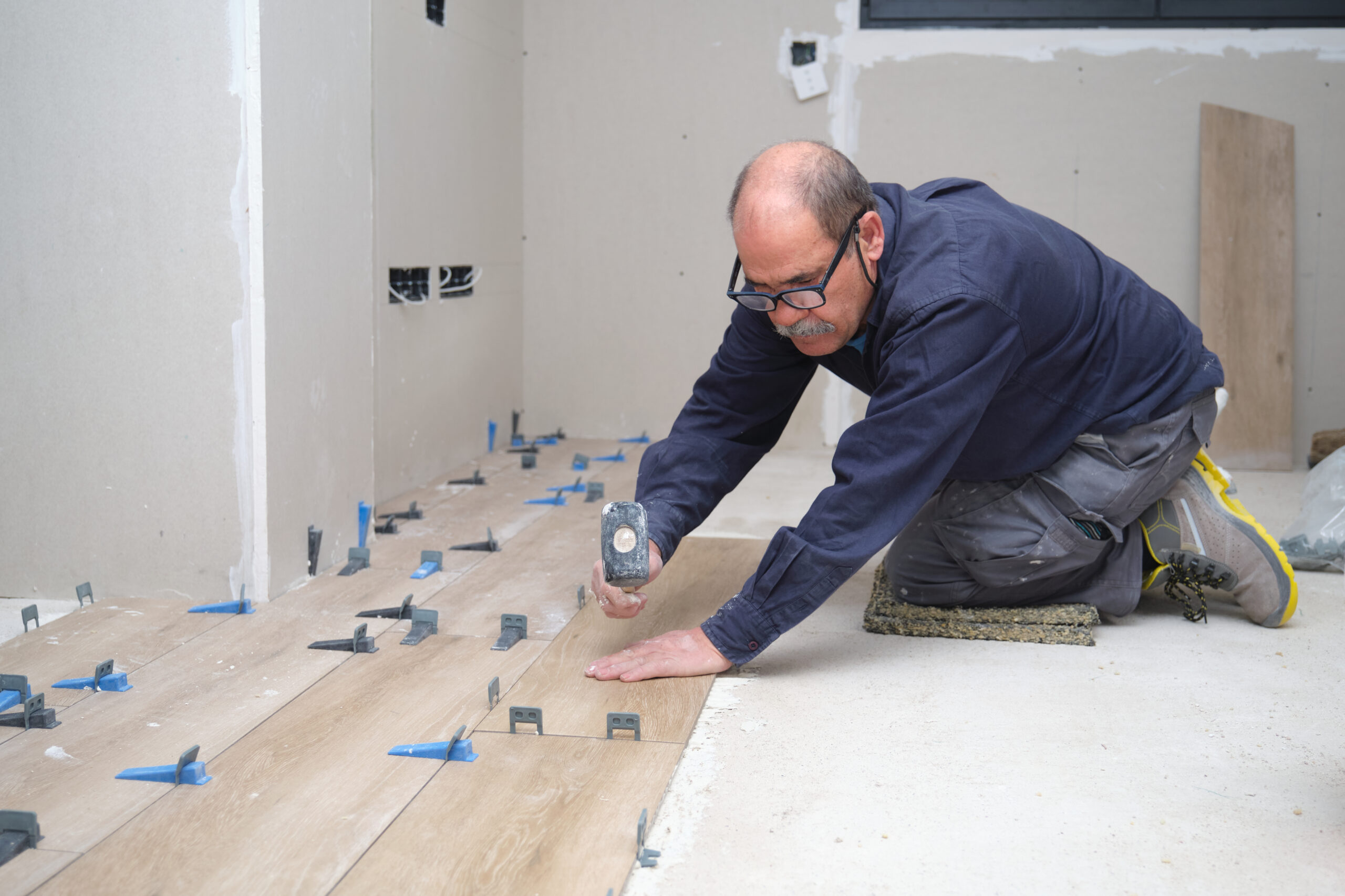 Senior contractor laying ceramic wood effect tiles on the floor using a mallet. House renovation.